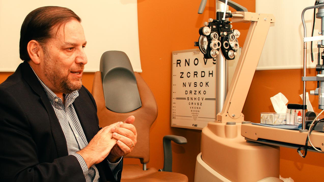 Doctor de oftalmología Jorge Eugenio Valdez García durante entrevista en su consultorio. Se observa diferentes elementos médicos como máquina de optometría, tabla de Snellen y una silla, entre otros