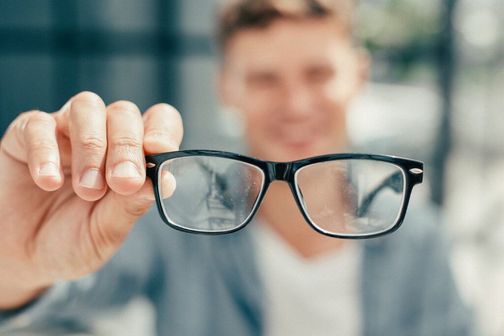 Hombre joven sosteniendo sus lentes ópticos hacia el frente, con desenfoque en el fondo de la imagen, relacionado a los beneficios del ejercicio en la salud visual