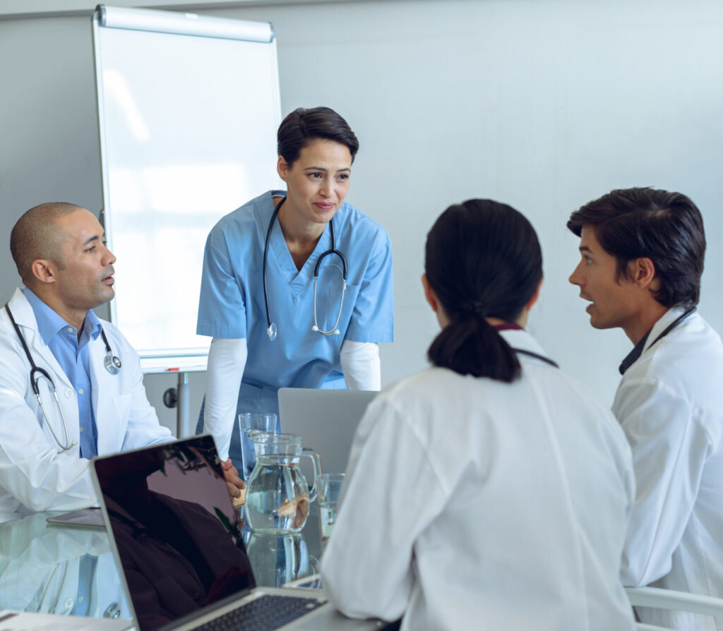 Vista frontal de una enfermera caucásica de pie atenta a un equipo médico diverso que habla entre sí y está sentada en la mesa de la sala de conferencias en el hospital.