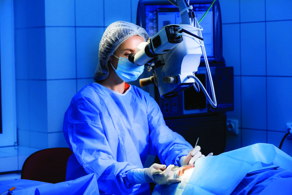 Una cirujana está operando en una sala quirúrgica. La mujer está mirando en el microscopio. La enfermera está ajustando una máscara estéril.