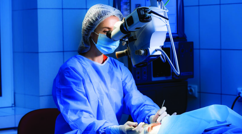 Una cirujana está operando en una sala quirúrgica. La mujer está mirando en el microscopio. La enfermera está ajustando una máscara estéril.